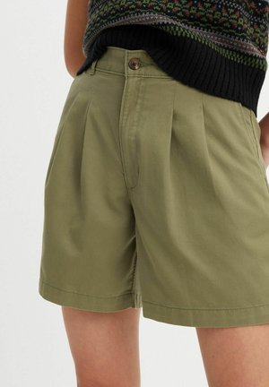 Femme portant un short plissé taille haute vert olive avec bouton et fermeture éclair à l'avant, associé à un haut sans manches en tricot à motifs.