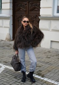 Bruine fuzzy jas, lichtblauwe jeans, zwarte veterschoenen en een donkere handtas in een straatsetting met een van klinkers gemaakt pad en een houten deur op de achtergrond.