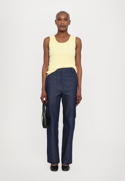 Gelbes, geripptes Tanktop mit gewellten Kanten, kombiniert mit dunkelblauen, weiten Jeanshosen mit Fronttaschen. Schwarze flache Schuhe und eine grüne Tasche.