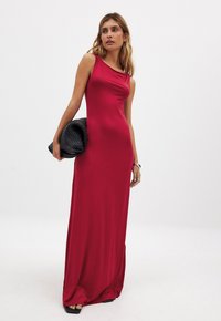 Rotes, ärmelloses, bodenlanges Kleid aus glattem Stoff mit einem dezenten Drapierungsausschnitt. Kombiniert mit einer strukturierten schwarzen Clutch.
