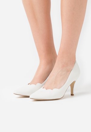 Des pieds chaussés de pompes à talons moyens à bout pointu et bords festonnés blancs sur un fond blanc uni.