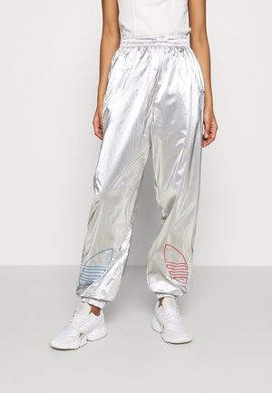 Personne portant un pantalon de survêtement argenté brillant avec une broderie de logo bleue et rouge sur le bas des jambes et des baskets blanches, debout devant un fond uni.