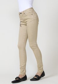 Vaqueros skinny beige de cintura alta, con costuras decorativas en la parte delantera y acentos metálicos. Textura suave con un diseño ajustado.