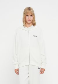 Bijela zip-up dukserica od rebraste tkanine sa slobodnim krojem, sa kapuljačom i prednjim džepovima. Crni "Pagator" logo na prsima.