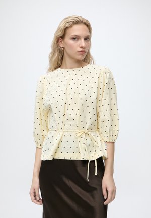 Vrouw met blond haar die een crèmekleurige blouse met zwarte stippen en een strik in de taille draagt, gecombineerd met een zwarte rok, staand tegen een effen achtergrond.