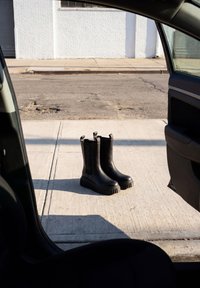 Paire de bottes noires épaisses posées debout sur un trottoir en béton, vue à travers une portière de voiture ouverte.
