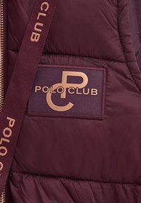 Vinrött quiltat tyg med en rektangulär "Polo Club" logotyp-patch och matchande vinrött band med samma text i beige bokstäver.