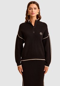 Czarny sweter z dzianiny, z wysokim kołnierzem, zapięciem na guziki oraz białymi akcentami na rękawach i dolnym brzegu. Posiada naszywkę z logo na piersi.