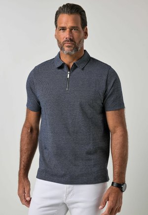 Mann mit Bart, der ein kurzärmeliges, marineblau gemustertes Zip-Kragen-Hemd und weiße Hose trägt, stehend vor einem einfachen hellen Hintergrund.