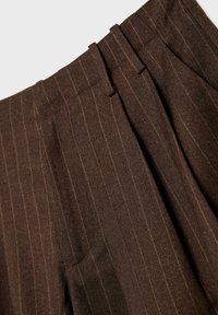 Detailný záber hnedých nohavíc s prúžkami, ktoré majú pútka na opasok a prehyby, zobrazuje textúru a vzor látky.