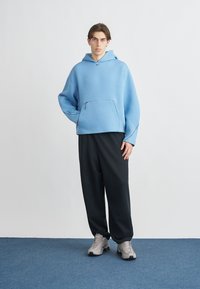 Light blue hooded sweatshirt with a front pocket, paired with black loose-fitting pants and grey sneakers. Smooth texture, relaxed fit.
