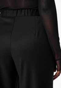 Zwarte broeken van gladde stof, met een relaxte pasvorm, elastische tailleband en zijzakken. De textuur lijkt zacht en mat.