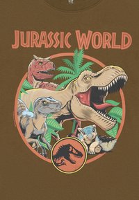 Ruda marškinėliai su apvalia grafika, vaizduojančia iliustruotus dinozaurus, įskaitant T. rex ir raptorą, apsuptus žalių lapų ir „Jurassic World“ pavadinimu.