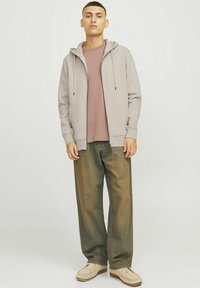 Hanorac cu fermoar de culoare gri deschis peste un tricou roz, asortat cu blugi verzi largi și pantofi slip-on bej. Materialul este neted, având o textură subtilă.