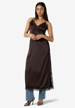 Jeune femme portant une robe nuisette en satin marron foncé avec bordure en dentelle sur un jean bleu et des sandales à talons, debout les mains jointes.
