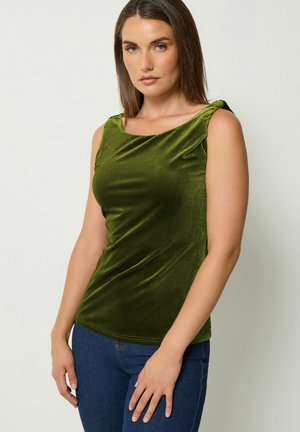 Donna con capelli castani lisci indossa un top verde oliva in velluto senza maniche e jeans blu, in piedi davanti a uno sfondo chiaro.