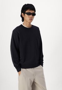 Sort crewneck sweatshirt med lange ærmer, ribbede manchetter og en glat tekstur. Bæret over en hvid skjorte, parret med lysfarvede bukser.