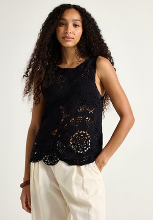 Ung kvinde med krøllet hår iført en ærmeløs sort hæklet top og løse hvide bukser, hånd i lommen, neutral baggrund.