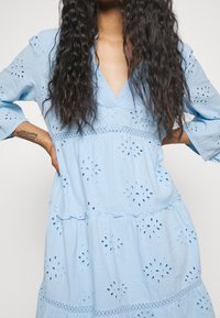 Ljust blå, lager-på-lager klänning med v-ringning, ögletmönster och volangärmar. Tillverkad av strukturerad tyg med dekorativa spetsdetaljer.