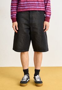Shorts en denim noir avec une coupe décontractée, comportant cinq poches et des coutures contrastées, associés à des baskets noires et des chaussettes noires.
