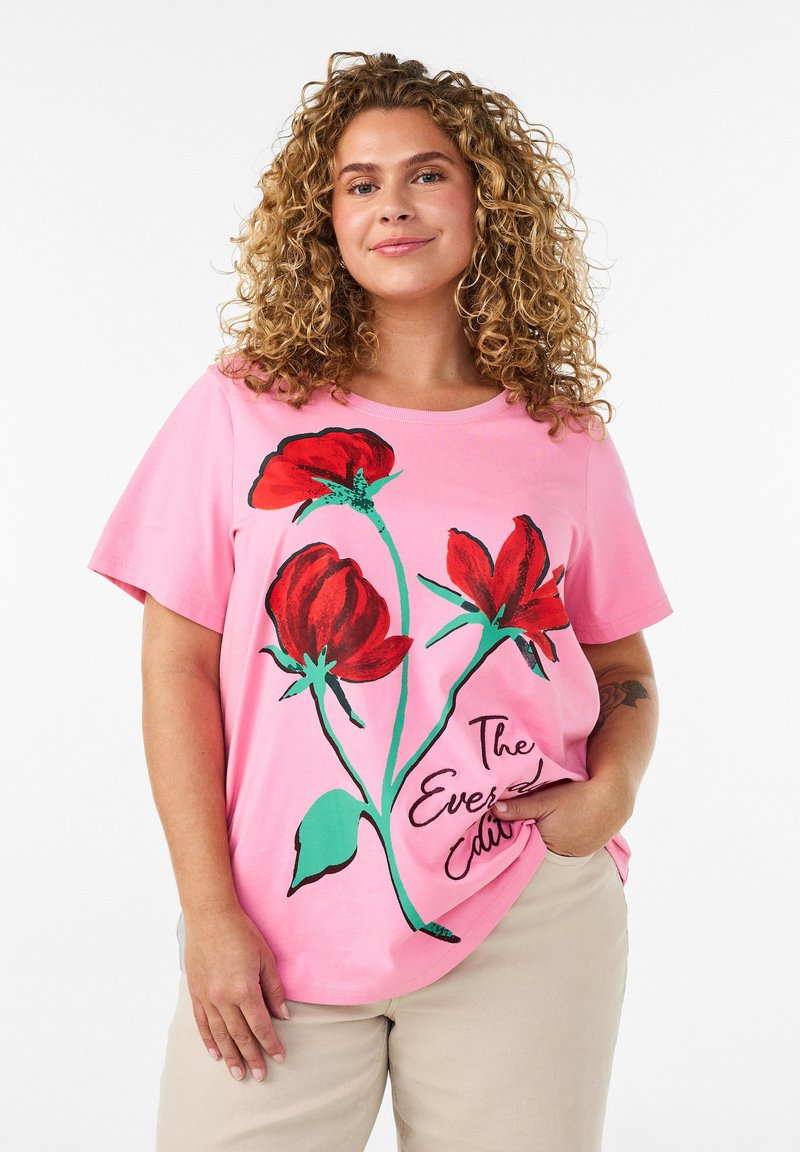 Femme aux cheveux bouclés portant un t-shirt rose avec de grandes fleurs rouges et un texte, pantalon beige, debout avec une main dans la poche.