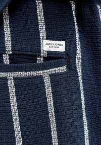Marineblåt tekstureret stof med hvide lodrette striber og et påsyet Jack & Jones-mærke med teksten "EST. 1990" nær en foldet kant.