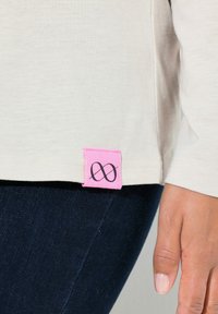 Helles beiges Langarmshirt mit einem pinken Stofflabel, das am Saum ein schwarzes Unendlichkeitssymbol zeigt. Weicher Stoff, figurbetontes Design.