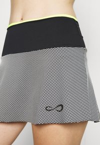 Falda de tenis con patrón en blanco y negro, con una cintura sólida negra que presenta un acento amarillo. El material parece elástico y ligero.