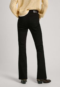 Jeans negros acampanados con una textura suave y bolsillos traseros, combinados con un suéter ligero de punto amarillo. El calzado presenta un tacón bajo.