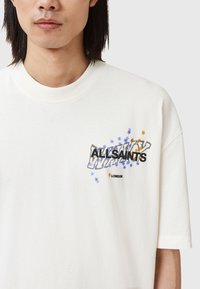 Camiseta de algodón blanca con un diseño gráfico que presenta el texto "ALLSAINTS", acentos de estrellas azules y naranjas dispersos, y un cuello redondo.