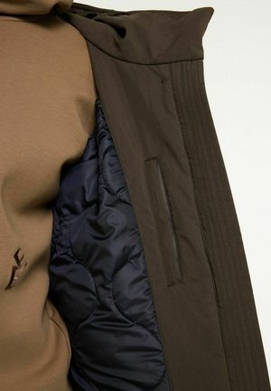 Brown outer fabric jacket with a navy quilted lining. Features a small zip pocket and textured detailing along the seams.
