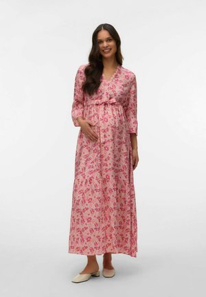 Femme enceinte souriante portant une longue robe rose à fleurs avec un nœud à la taille, debout pieds nus sur un fond blanc uni.