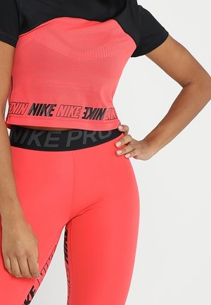 Vrouw draagt fel koraalkleurige Nike sportlegging en bijpassende crop top met zwarte Nike-logo banden, hand op de heup.