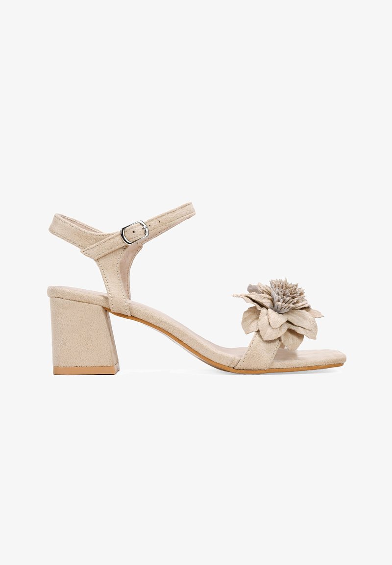 Sandalias de tacón de ante beige con tacón bloque, correa de tobillo ajustable y acento de flor de tela en la punta. Textura suave y diseño minimalista.