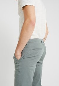 Pantaloni grigi chiari slim-fit realizzati in un tessuto misto cotone. Presentano tasche frontali, una tasca posteriore e una texture liscia.