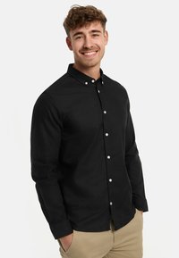 Mladý muž sa usmieva, má na sebe čiernu košeľu na gombíky a béžové nohavice, ruky má v kapse, stojí pred bielym pozadím.