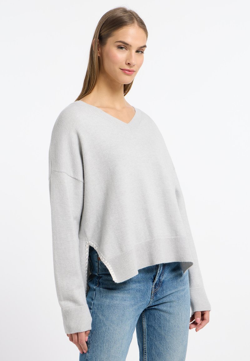Hellgrauer Strickpullover mit V-Ausschnitt, oversized geschnitten, mit gerippten Bündchen und seitlichen Schlitzen. Kombiniert mit blauen Jeans im lässigen Design.