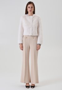 Weißes, kurzes Jacke aus Kunstpelz mit rundem Ausschnitt, kombiniert mit beigen, weit geschnittenen Hosen mit goldenen Knopfakzenten. Schwarze spitze Schuhe.