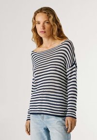 Mujer con cabello rubio ondulado que lleva un suéter a rayas marino y blancas con los hombros descubiertos y unos vaqueros azul claro, de pie contra un fondo liso.