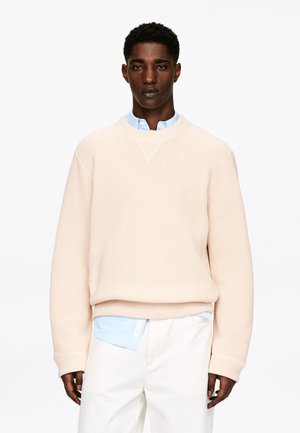 Hellrosa gestrickter Pullover mit Rundhalsausschnitt und Rippenstruktur. Darunter ein hellblaues Hemd mit Kragen, kombiniert mit weißen Hosen.