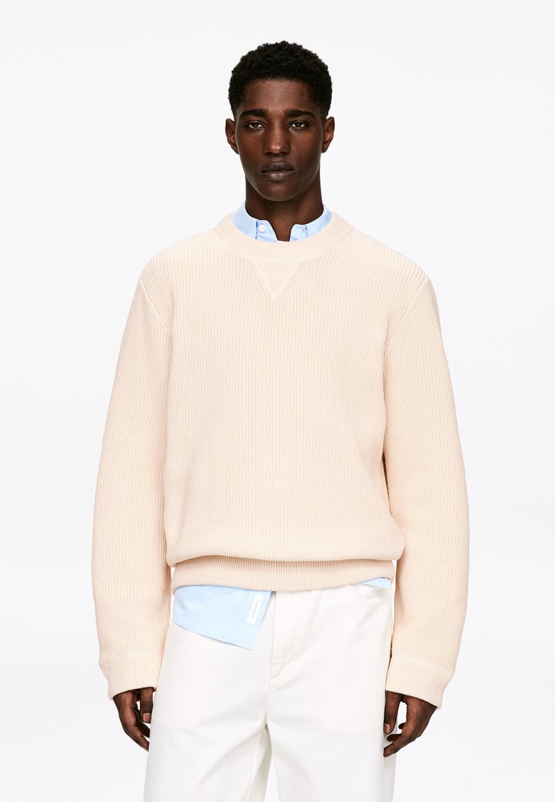 Pull rose clair en maille avec un col rond, présentant une texture côtelée. Porté par-dessus une chemise bleu clair à col, assorti à un pantalon blanc.