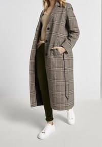 Karierter langer Mantel in Braun- und Beigetönen, mit maßgeschneiderter Passform, Knöpfen und seitlichen Taschen. Kombiniert mit grünen Hosen und weißen Sneakers.