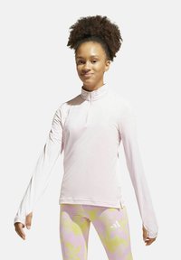 Maglietta sportiva a maniche lunghe di colore rosa chiaro con colletto a mezza zip. Texture liscia, design aderente. Indossata con leggings a pastello a fantasia.