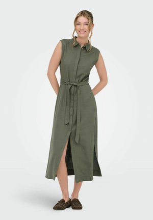 Naeratav naine, kandes varrukateta oliivrohelist vööd kinnitatavat midi kleiti esislõhikuga ja pruune libisevaid kingi, seisab ühtlase tausta ees.
