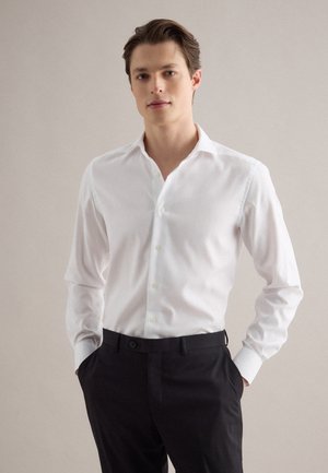 Junger Mann, der ein weißes Hemd mit Knöpfen und schwarze Hosen trägt, steht mit den Händen in den Taschen vor einem einfarbigen, hellen Hintergrund.