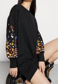 El vestido negro de algodón tiene mangas largas con colorido bordado floral en los puños. La tela es suave y tiene una silueta relajada.