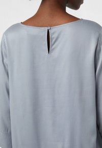 Blusa grigia chiaro con una texture liscia, dotata di scollatura tonda, maniche a tre quarti e un dettaglio a chiave sul retro fissato da un bottone.