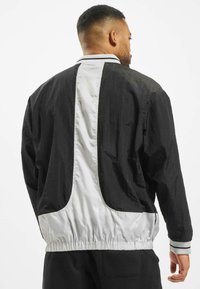 Chaqueta ligera en blanco y negro con cintura ajustable mediante cordón, que presenta una textura suave y un diseño de panel trasero en contraste.