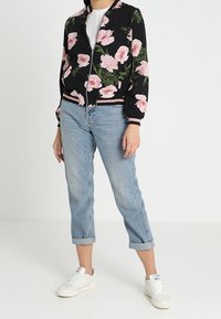 Giacca bomber nera con ampio motivo floreale rosa, polsini e orlo a costine, abbinata a jeans in denim azzurro arrotolati e sneakers bianche.