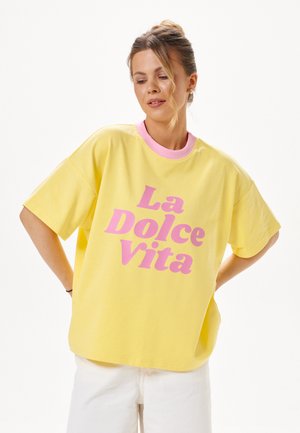 Kobieta ubrana w luźną żółtą koszulkę z różowym kołnierzykiem i napisem "La Dolce Vita" w różowym kolorze, w połączeniu z białymi spodniami, patrząca w dół.